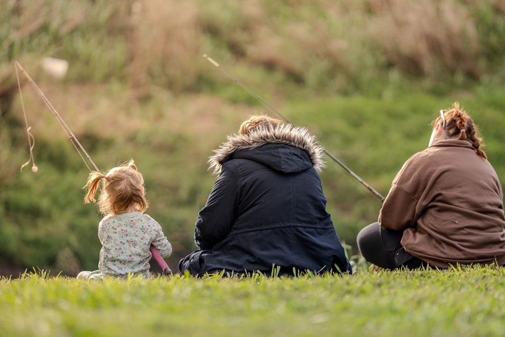 Three people sitting together on a grassy hill on a sunny day, viewed from behind.