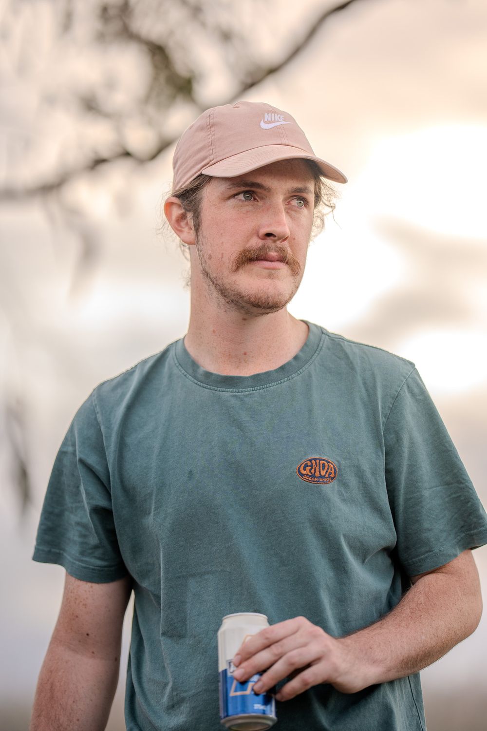 Person wearing a light pink Nike cap and green t-shirt holds a disposable cup while looking into the distance.