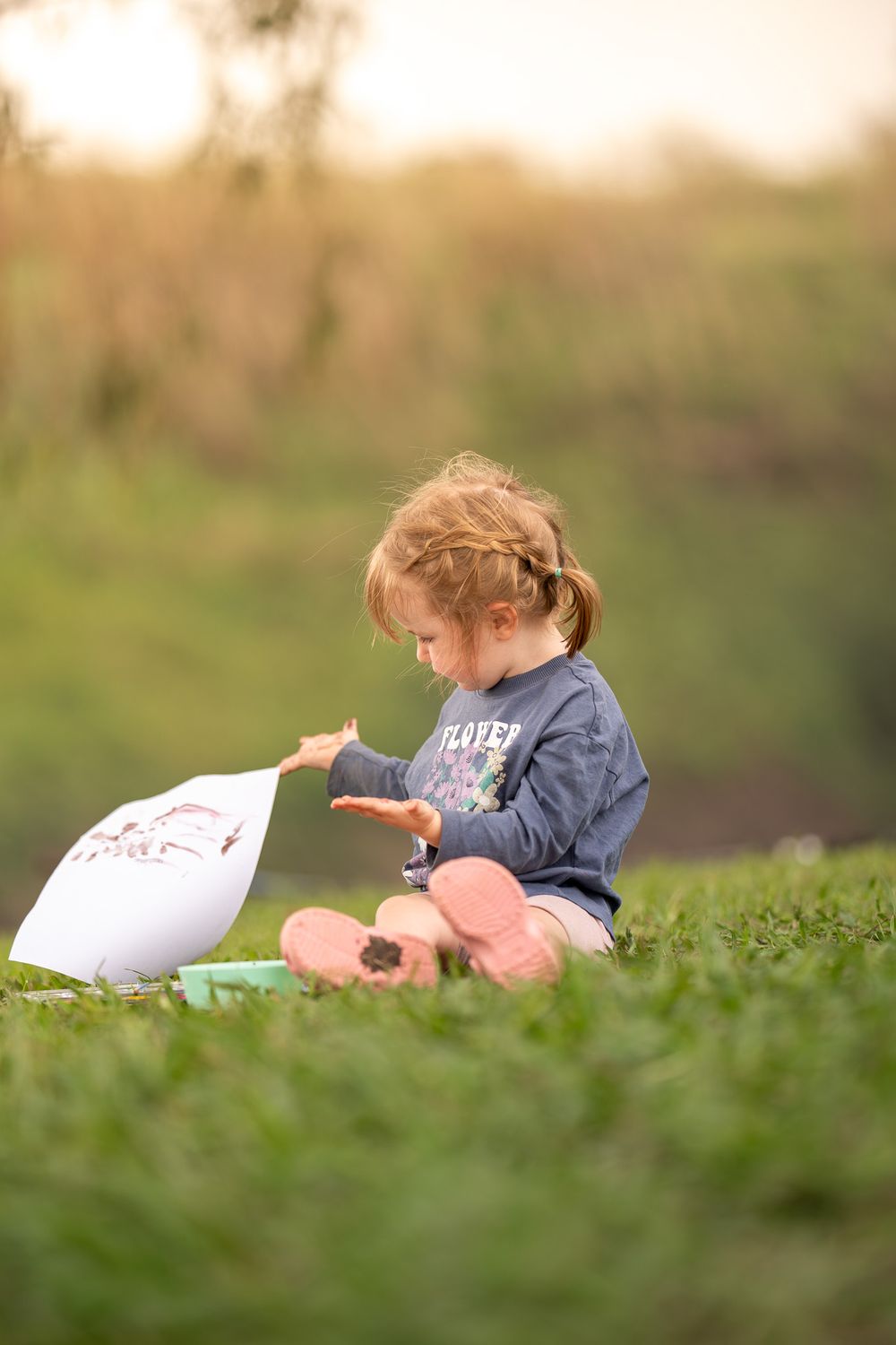 A small child in a denim shirt sits on grass at sunset while playing with a white object.