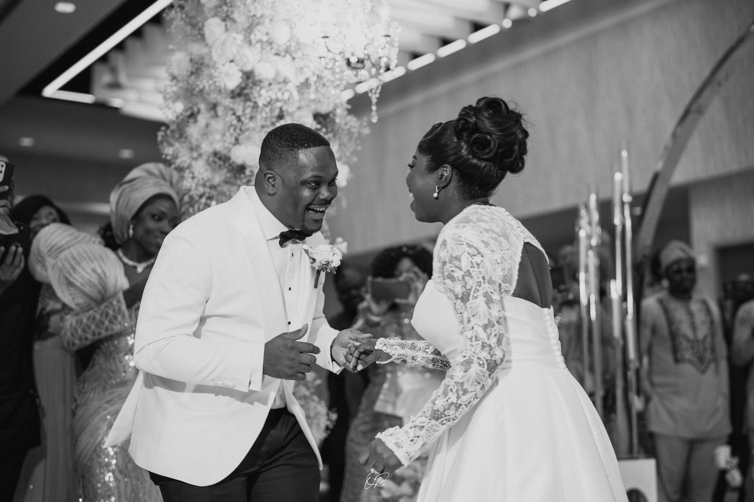 Black and white photo of elegant couple exchanging rings during wedding ceremony as guests look on in background.