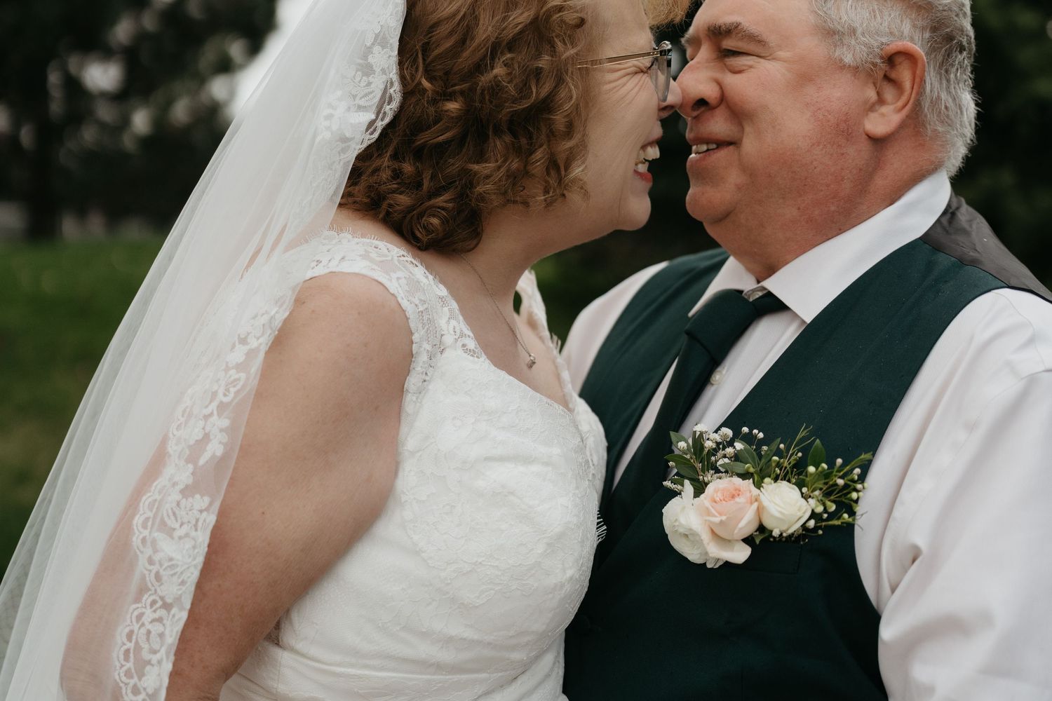 Sweet candid moment shared between a bride in white lace and a person wearing a dark vest at an outdoor wedding celebration.