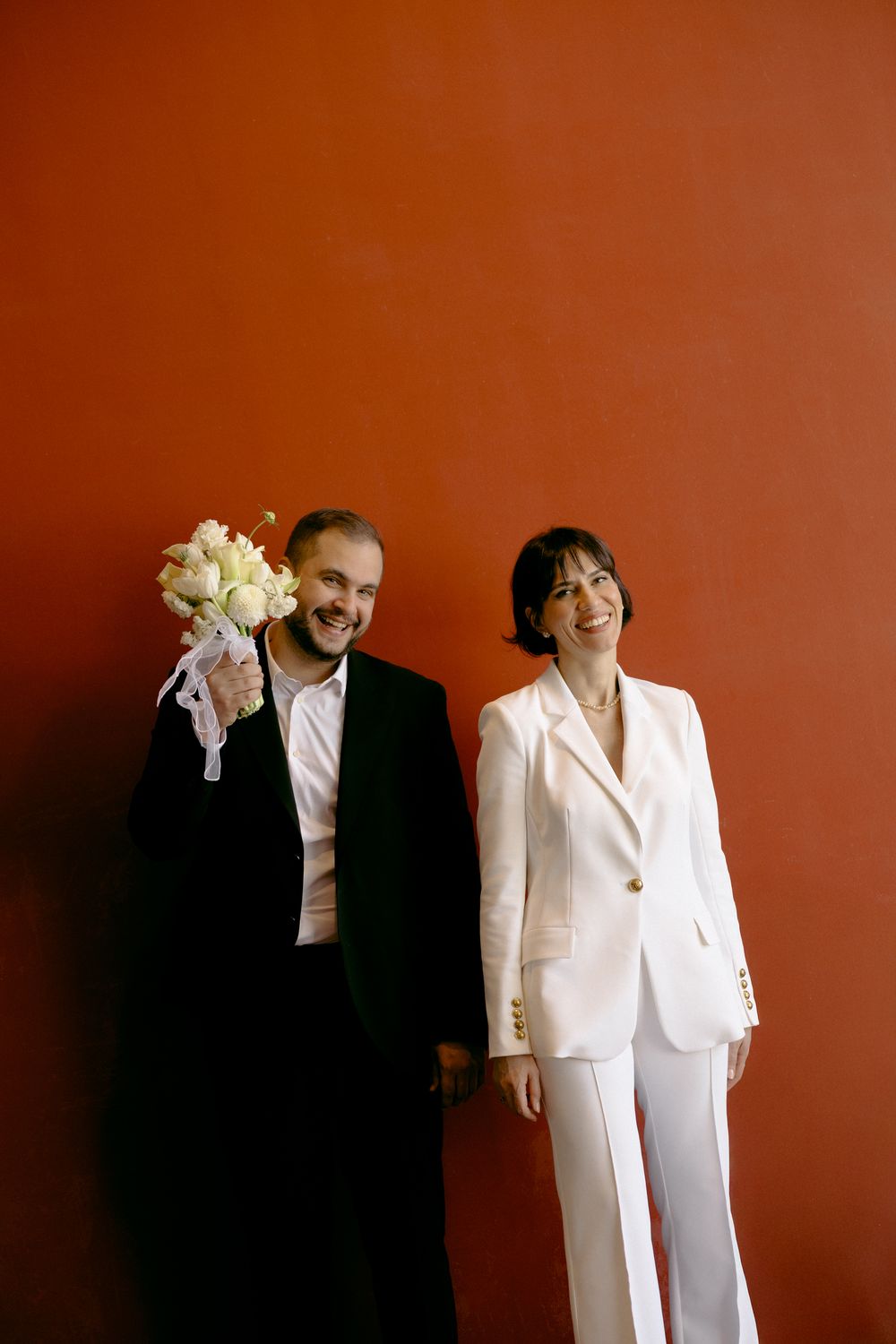 Wedding couple in black and white attire stand against vibrant red wall with bouquet. Destination wedding in London, UK.