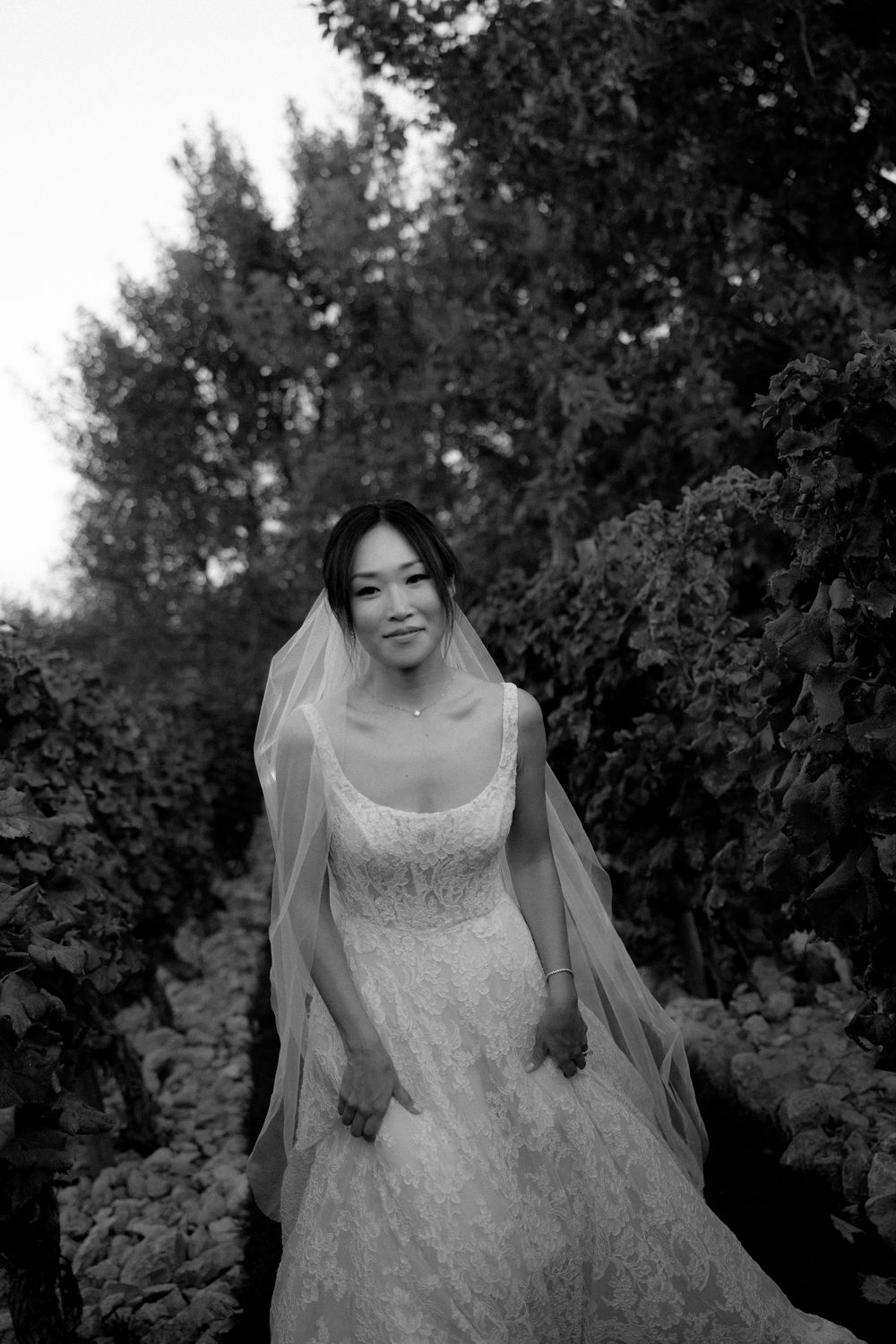 A bride in lace gown and veil stands among garden greenery in black and white portrait. Pyrgos Petreza, Athens.