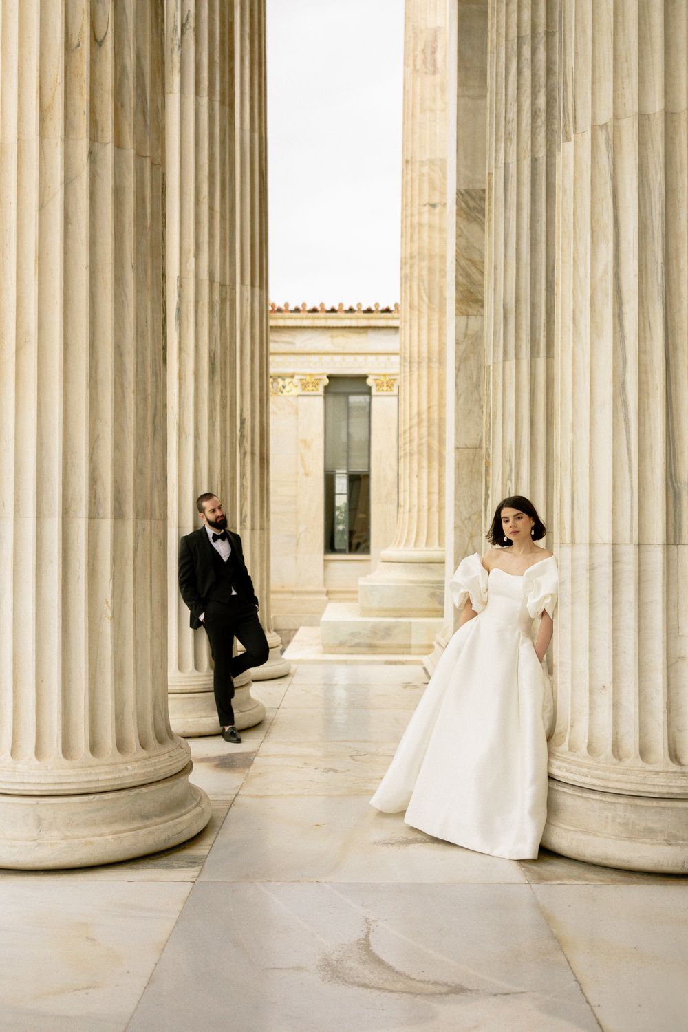 Stylish couple in wedding attire posed between towering classical columns. Athens, Greece.