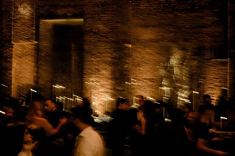 Warm lighting creates silhouettes of people gathering in a brick-walled venue. Block 33, Destination Wedding in Thessaloniki, Greece.