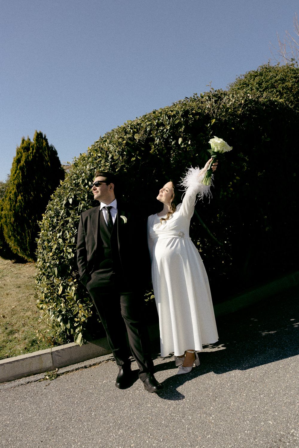 Stylish wedding couple poses playfully against lush garden hedge. Destination wedding in Greece.