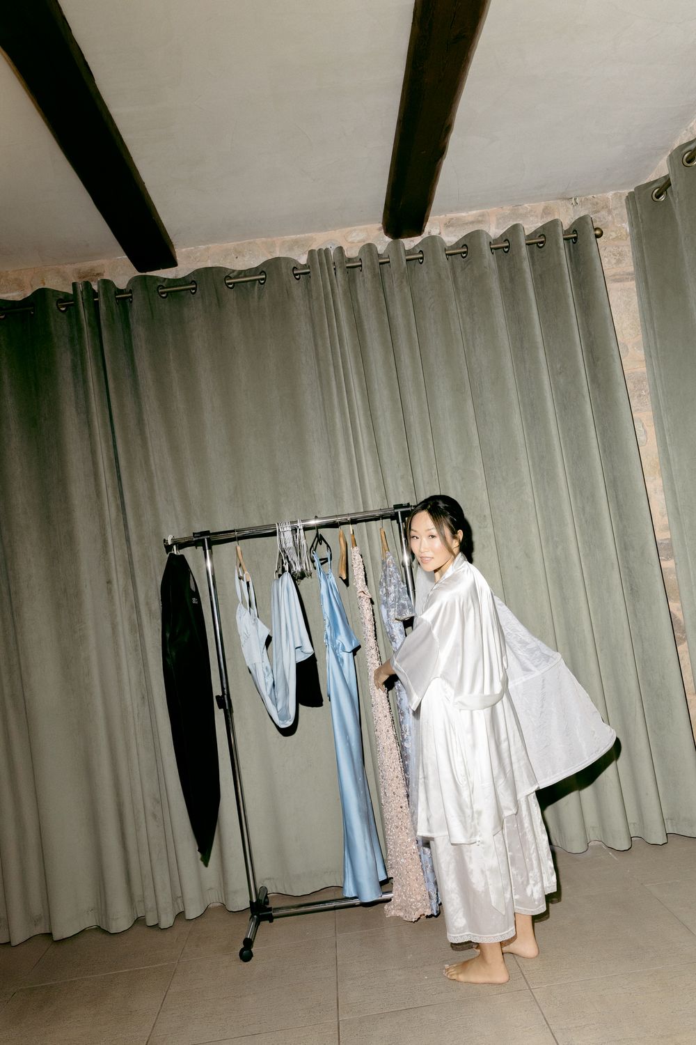 Person in white robe looking at hanging dresses against grey curtain background.