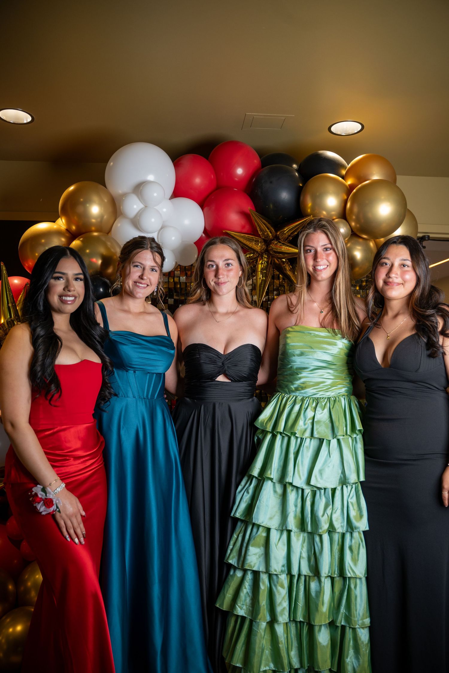 Group of friends in formal dresses posing together at party with balloon decorations.
