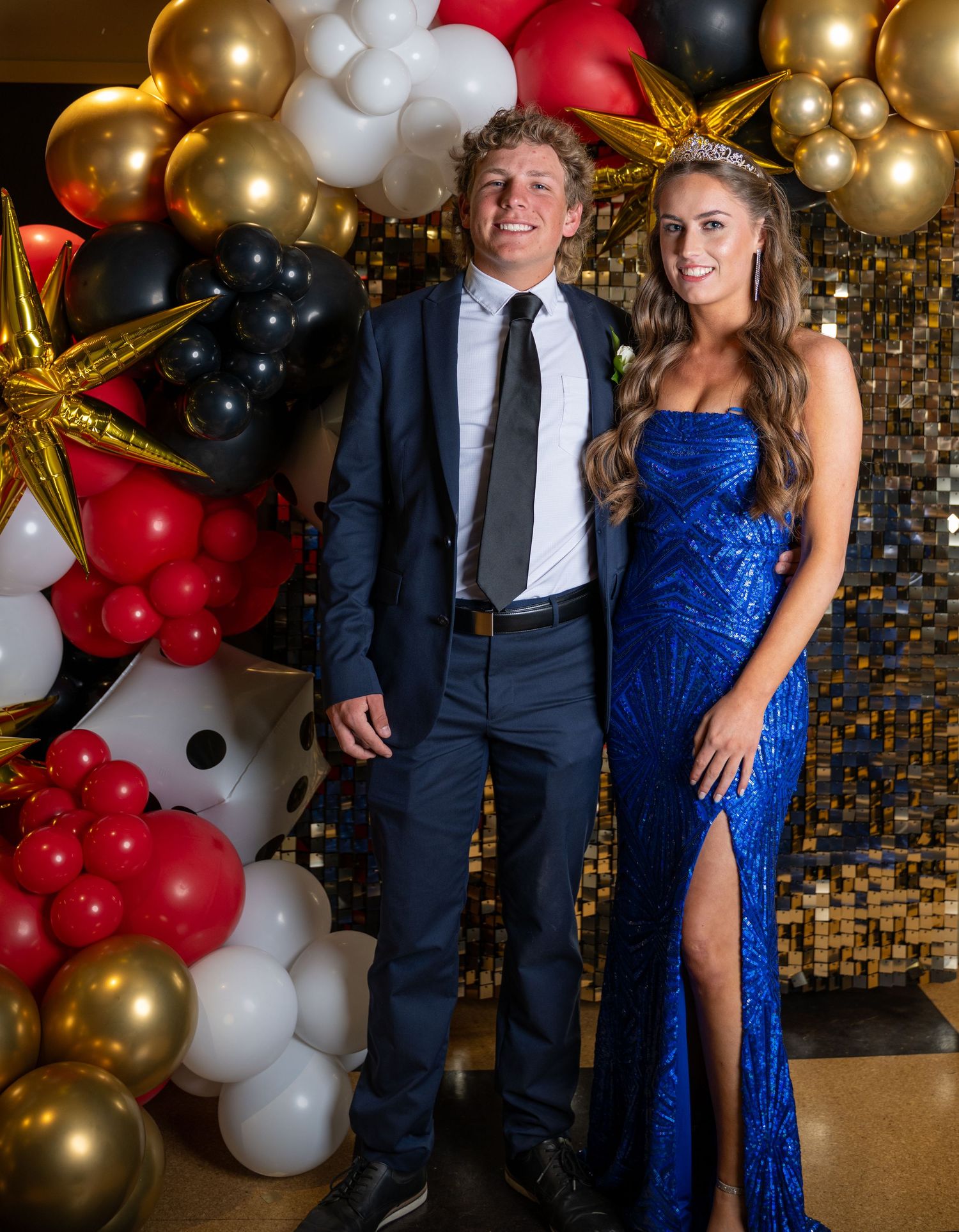 Couple in formal wear posing against festive balloon wall at special occasion.