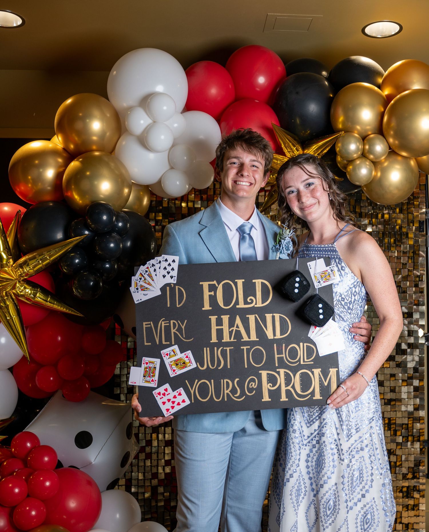 Decorative balloon arch with gold, white and red balloons frames a casino-themed prom proposal sign.