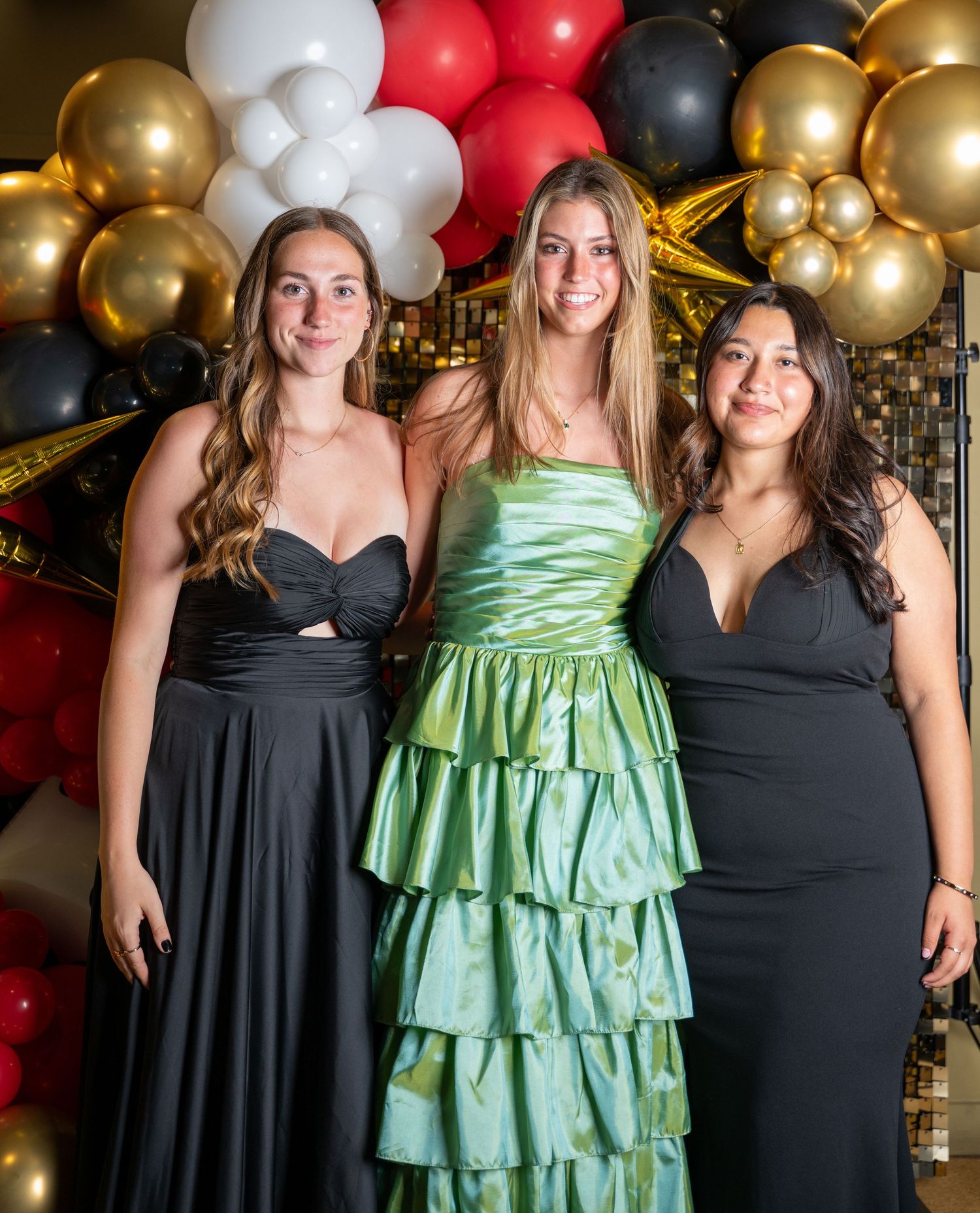 Three friends pose in formal dresses against a festive balloon backdrop at a special event.