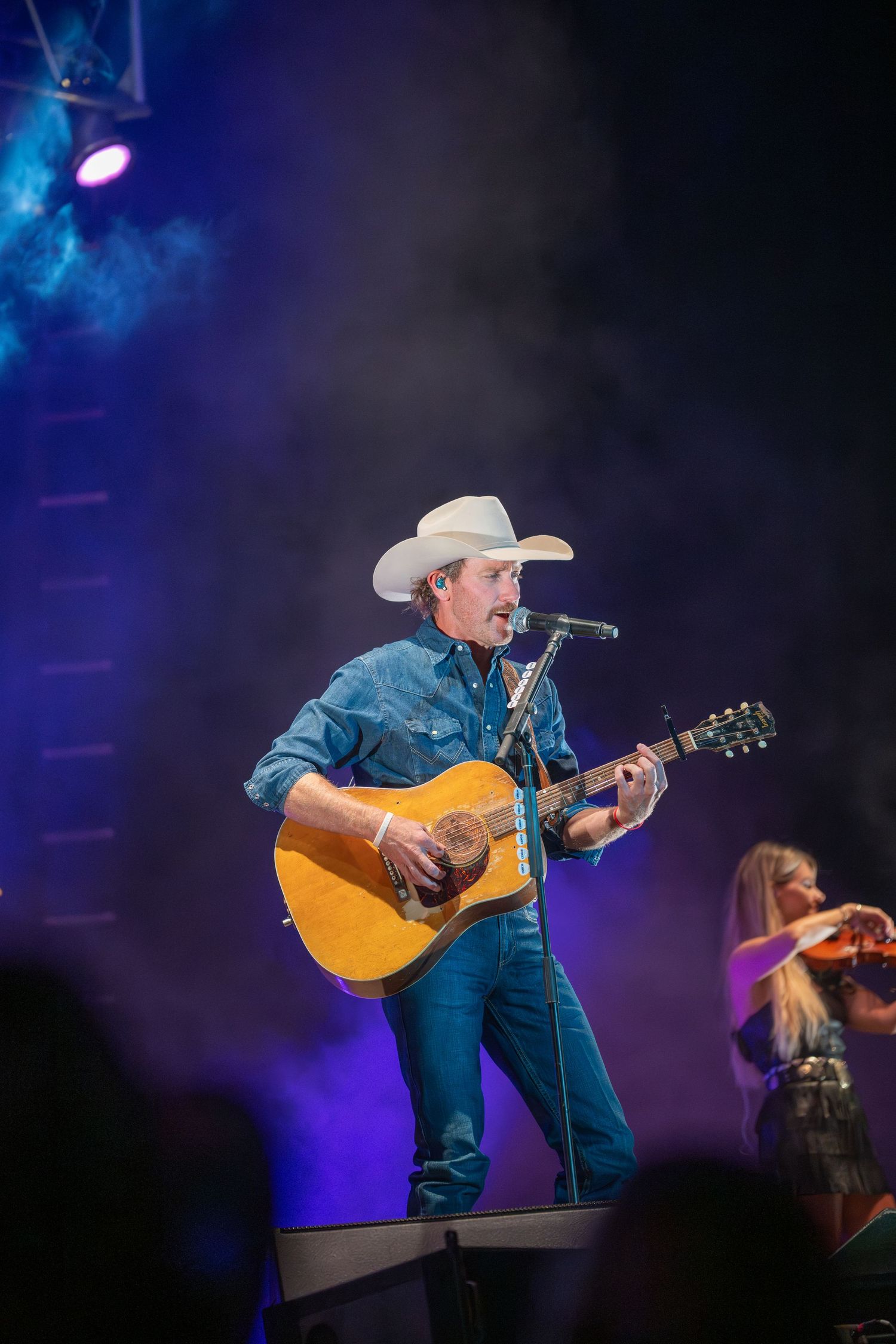 Country music performer plays acoustic guitar on stage with blue lighting and fog effects during live concert performance.