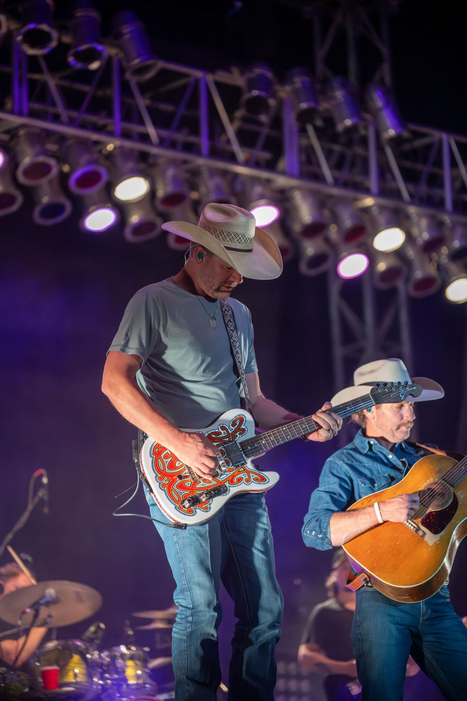 Musicians perform on stage with guitars and cowboy hats under purple stage lighting at a country music concert.