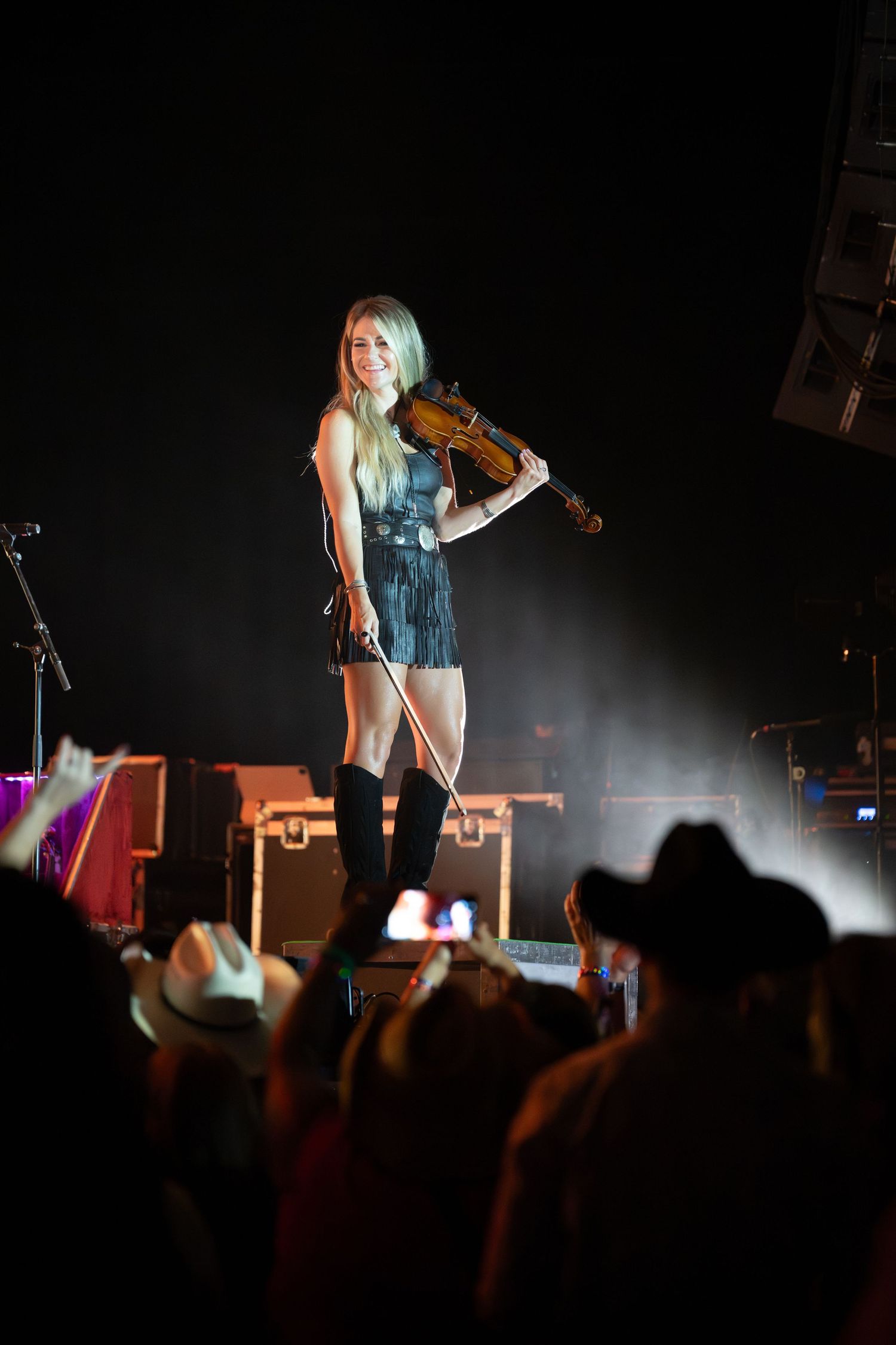 Performer in white outfit illuminated by stage lights while crowd silhouettes raise phones during concert performance.