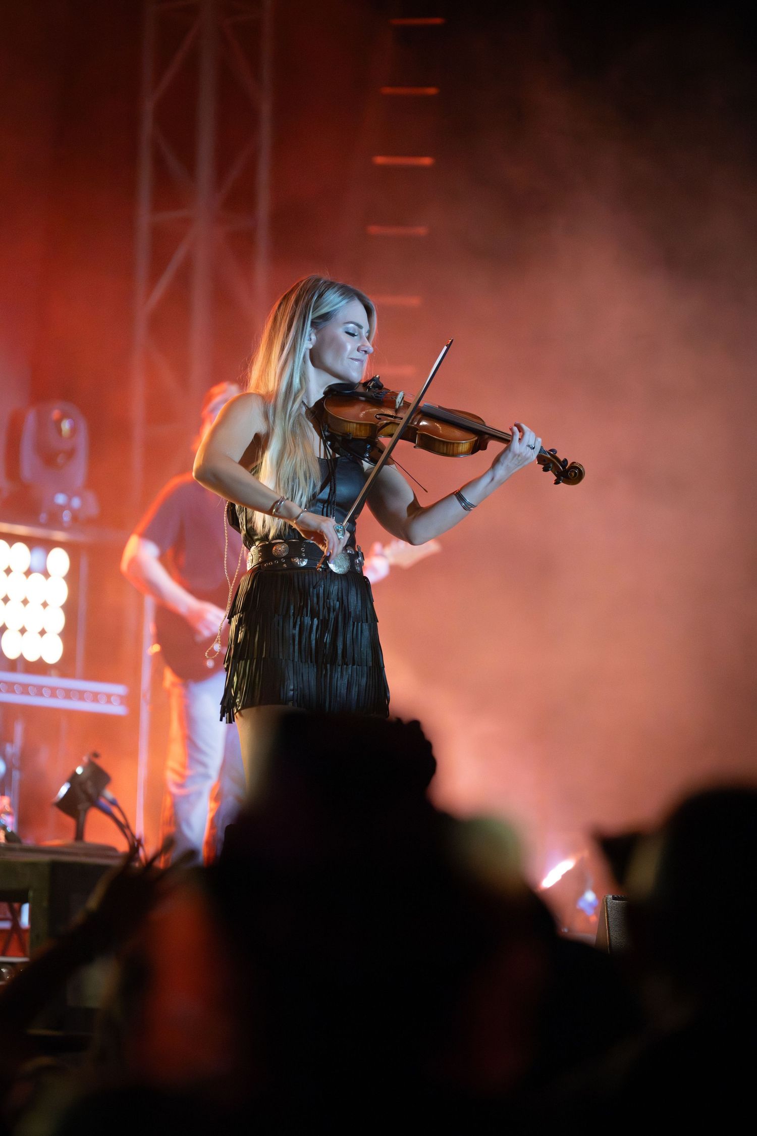 A violinist performs on stage bathed in dramatic red lighting while silhouetted audience members watch in the foreground.