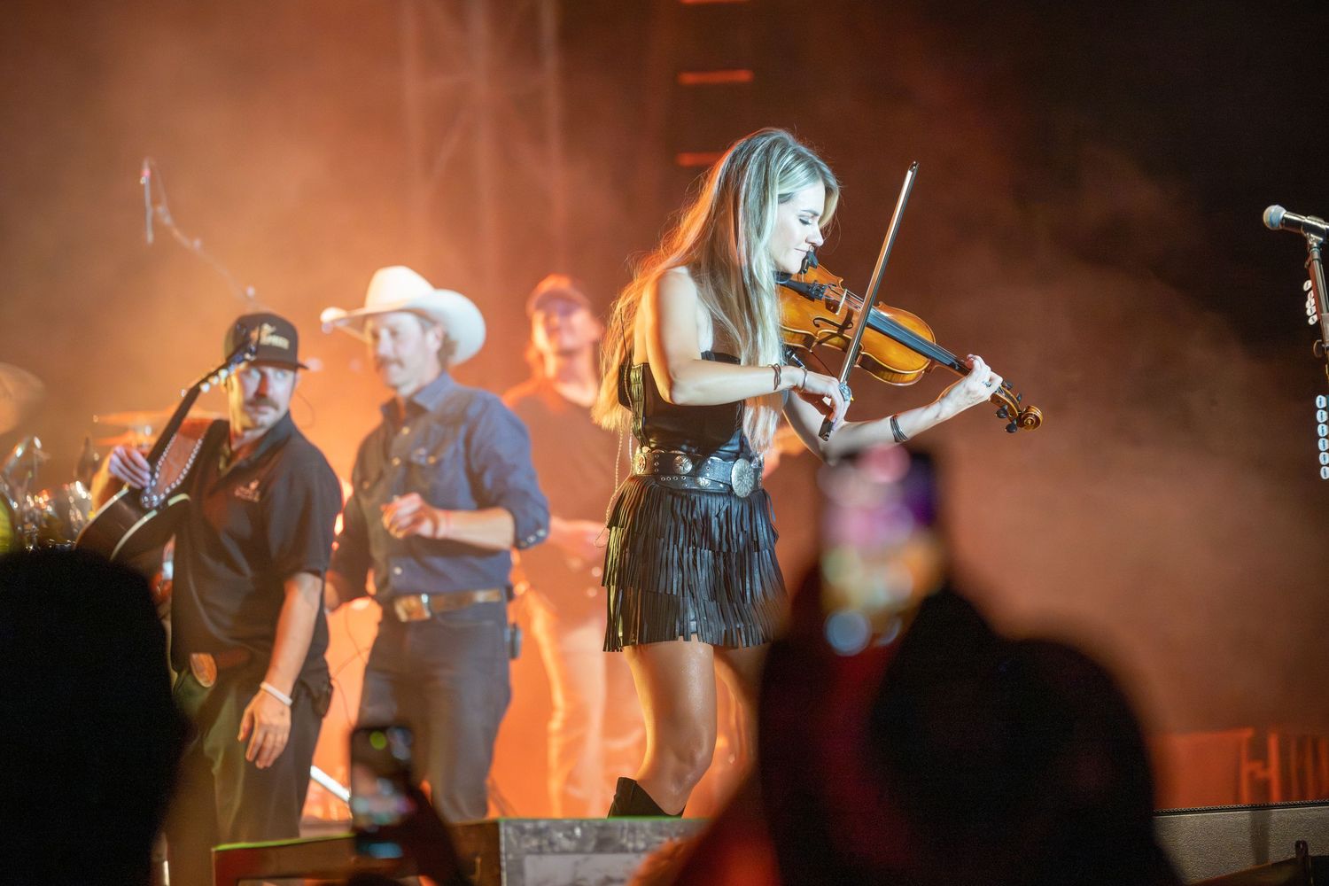 Musicians perform on stage with violins and instruments during a dynamic concert performance with dramatic lighting.