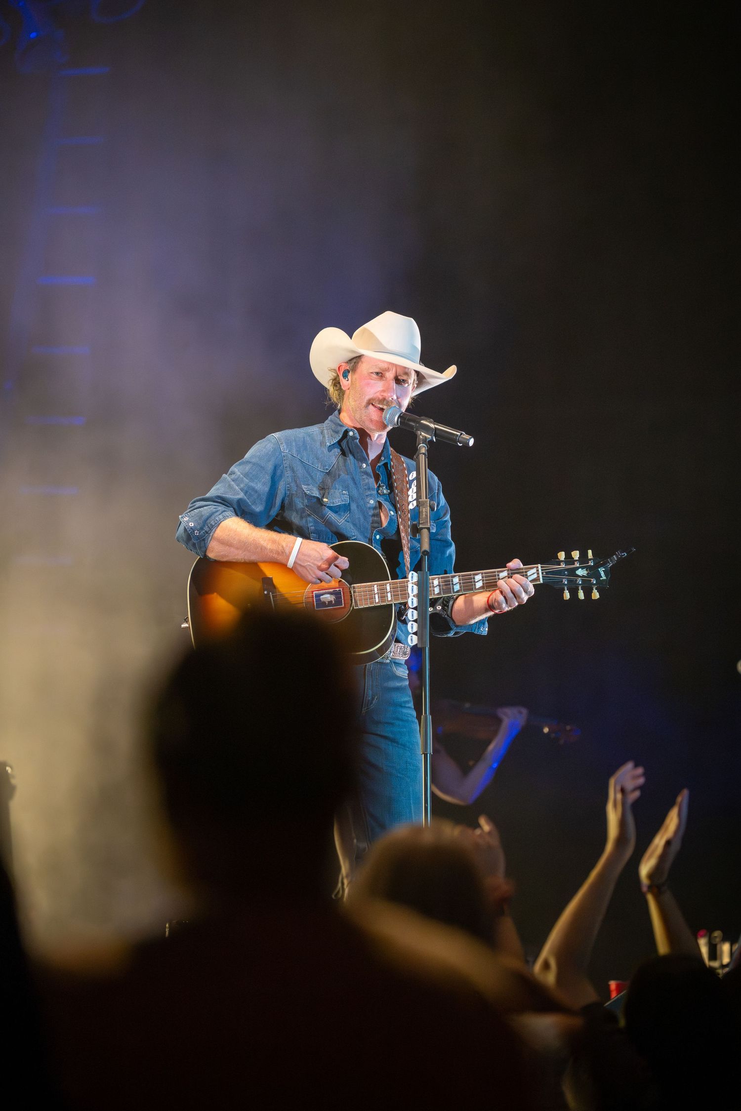 Country music performer in cowboy hat plays acoustic guitar on dark stage with silhouetted audience in foreground.