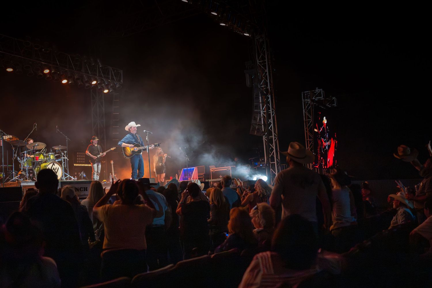 A dimly lit concert stage with performers silhouetted against colorful lighting and fog effects during a live performance.