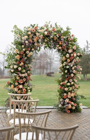 A floral wedding arch with peach and pink roses stands on a patio with wooden chairs.