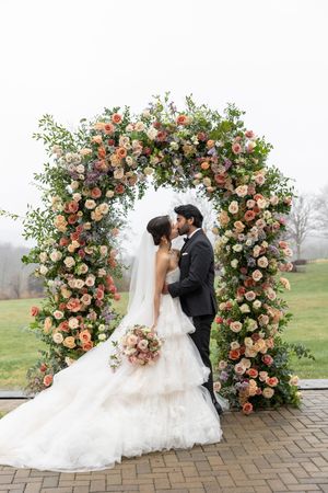 Romantic floral arch with pink and cream roses frames wedding ceremony on brick patio.