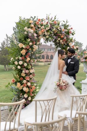 Wedding couple poses under beautiful peach floral arch with elegant venue in background.