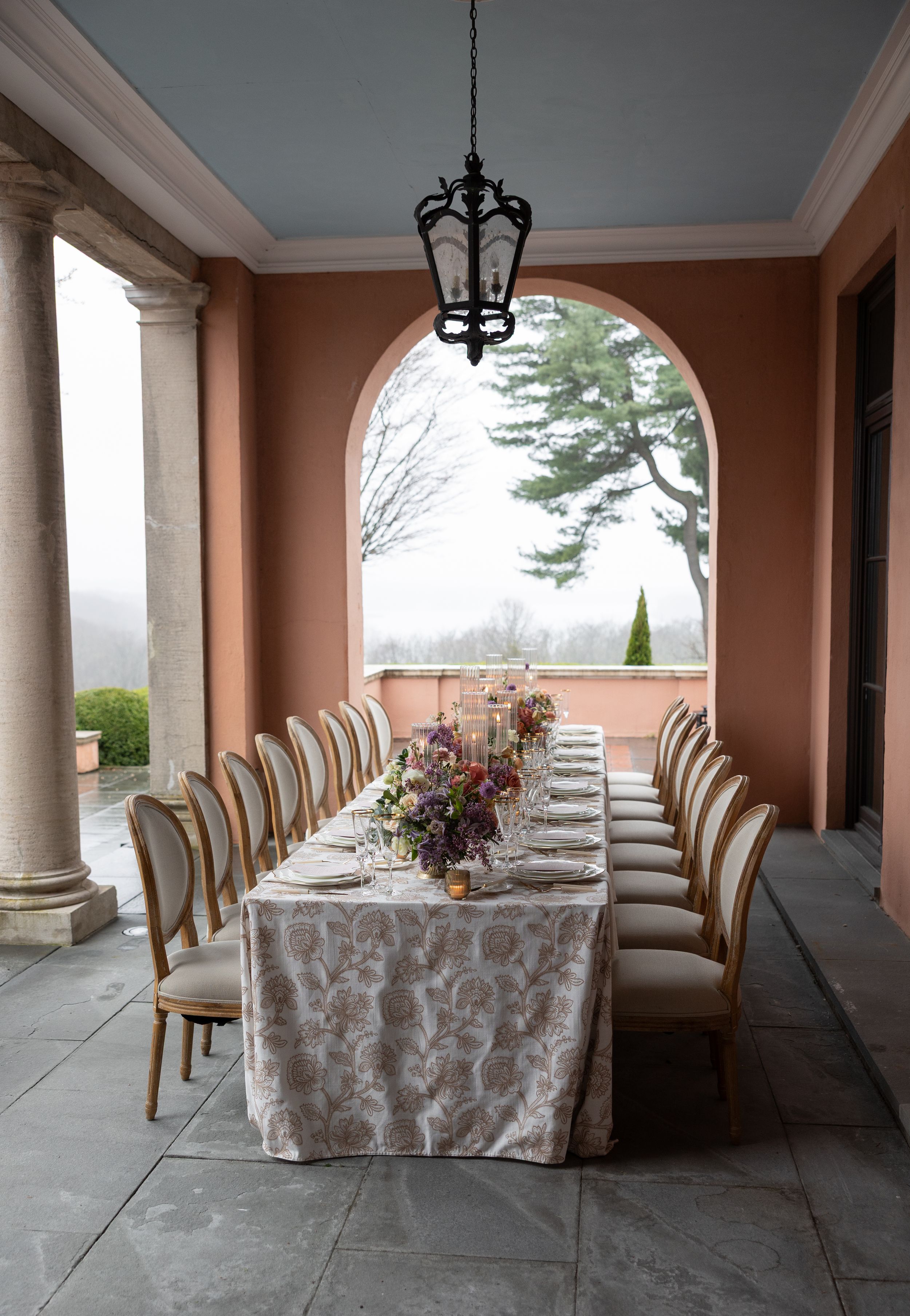Elegant outdoor dining table set under covered terrace with arched columns and wrought iron chandelier.