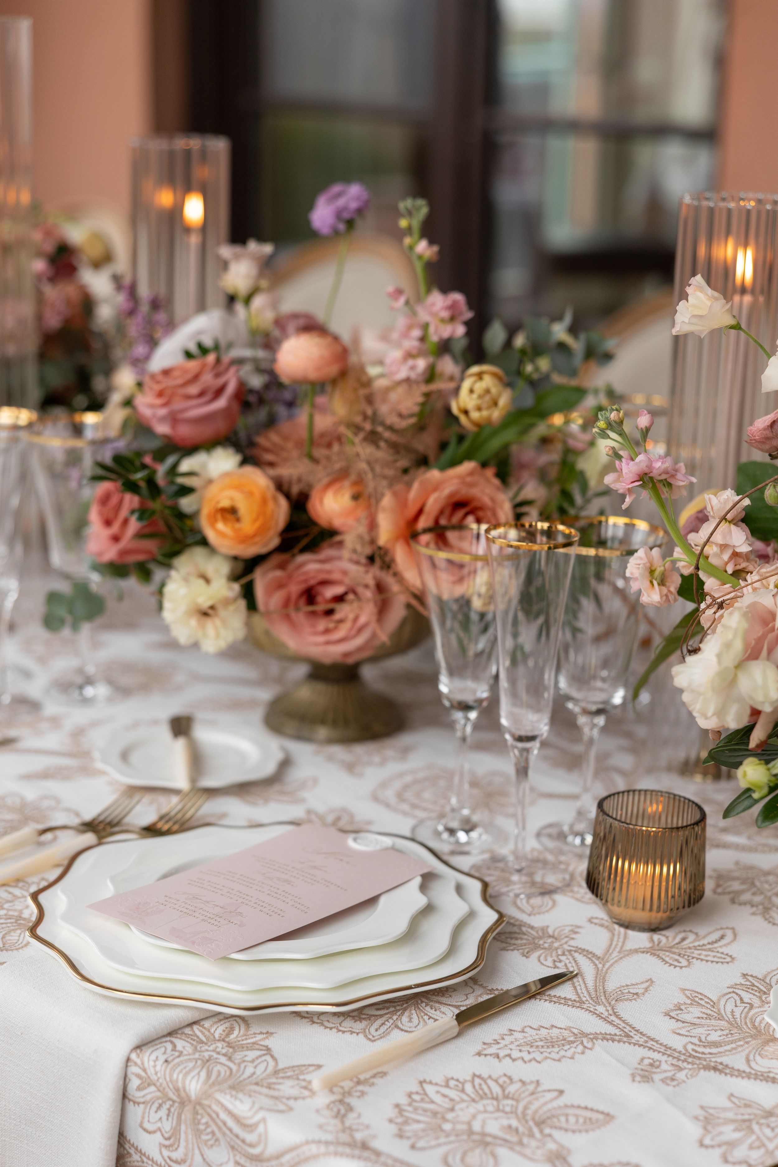 Elegant wedding table setting with peach and pink roses, white plates, gold flatware, and textured damask tablecloth.