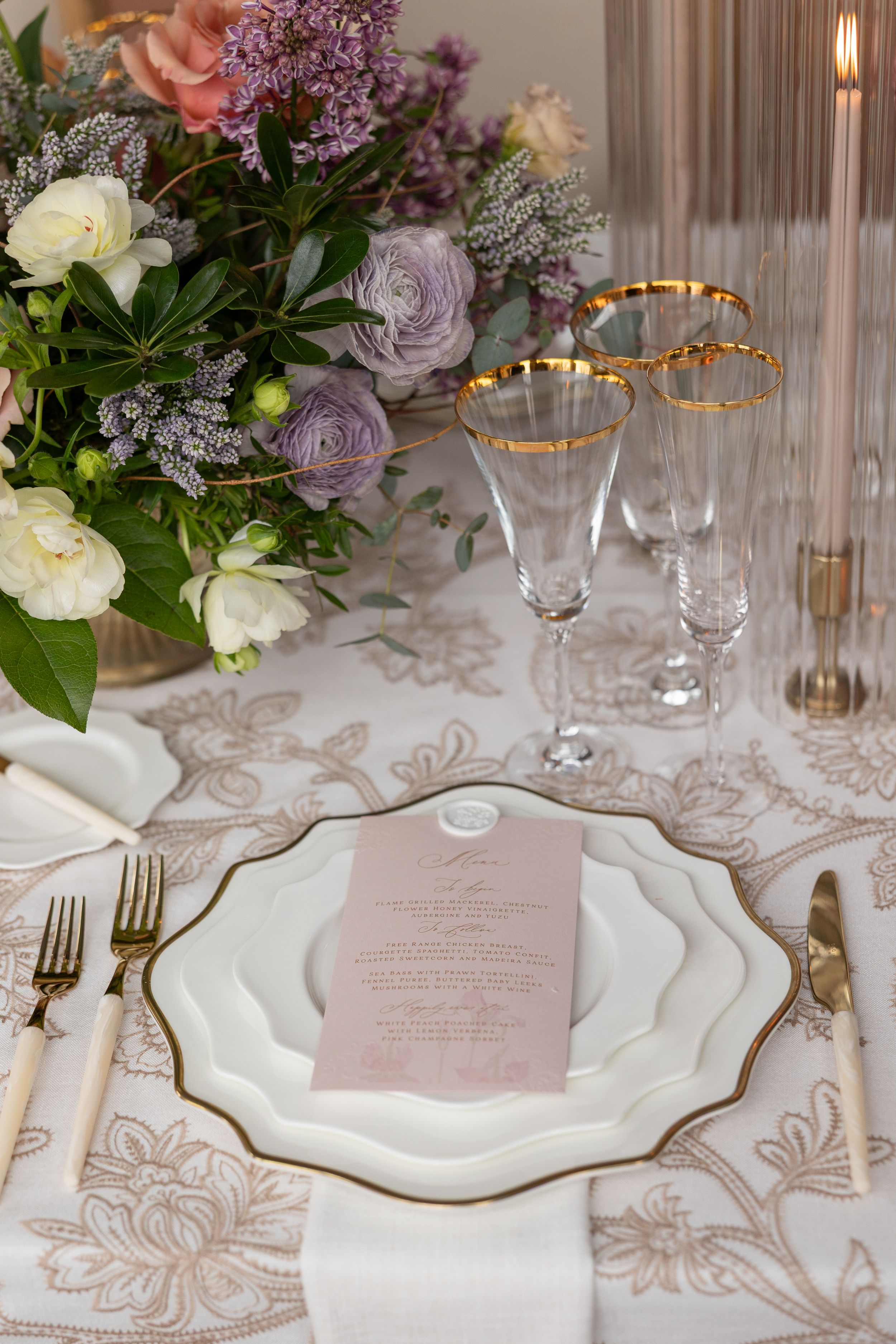 Elegant table setting with white plates, gold-rimmed glasses, and purple floral centerpiece.
