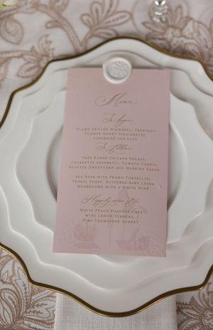 Blush pink wedding menu card with wax seal on scalloped white china plate.