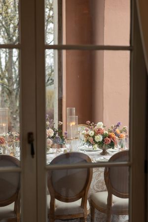 Elegant table setting viewed through French doors with floral centerpieces, candles and antique chairs in soft lighting.