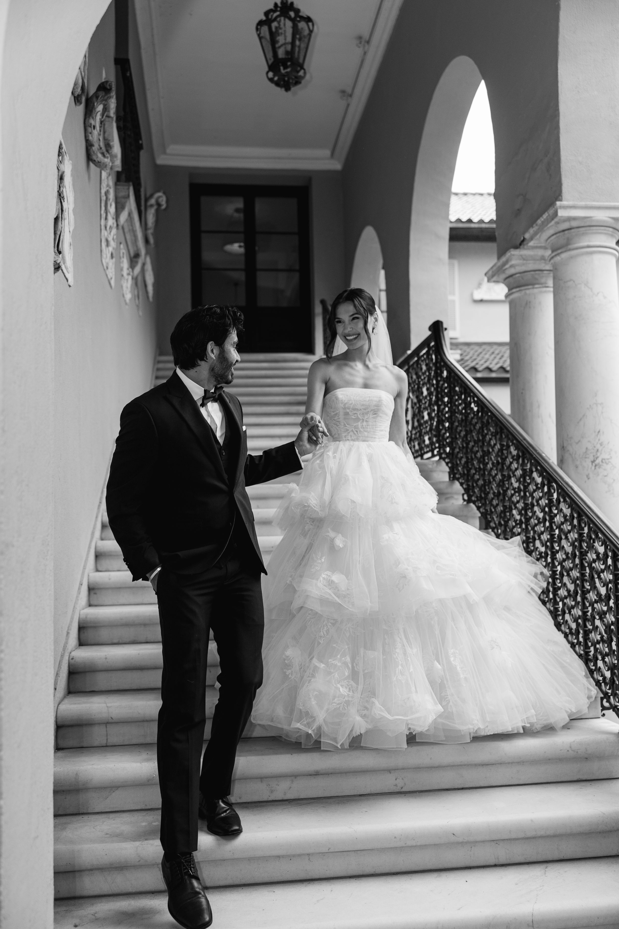 A couple in formal wedding attire descend an elegant marble staircase with ornate railings in a classic mansion.