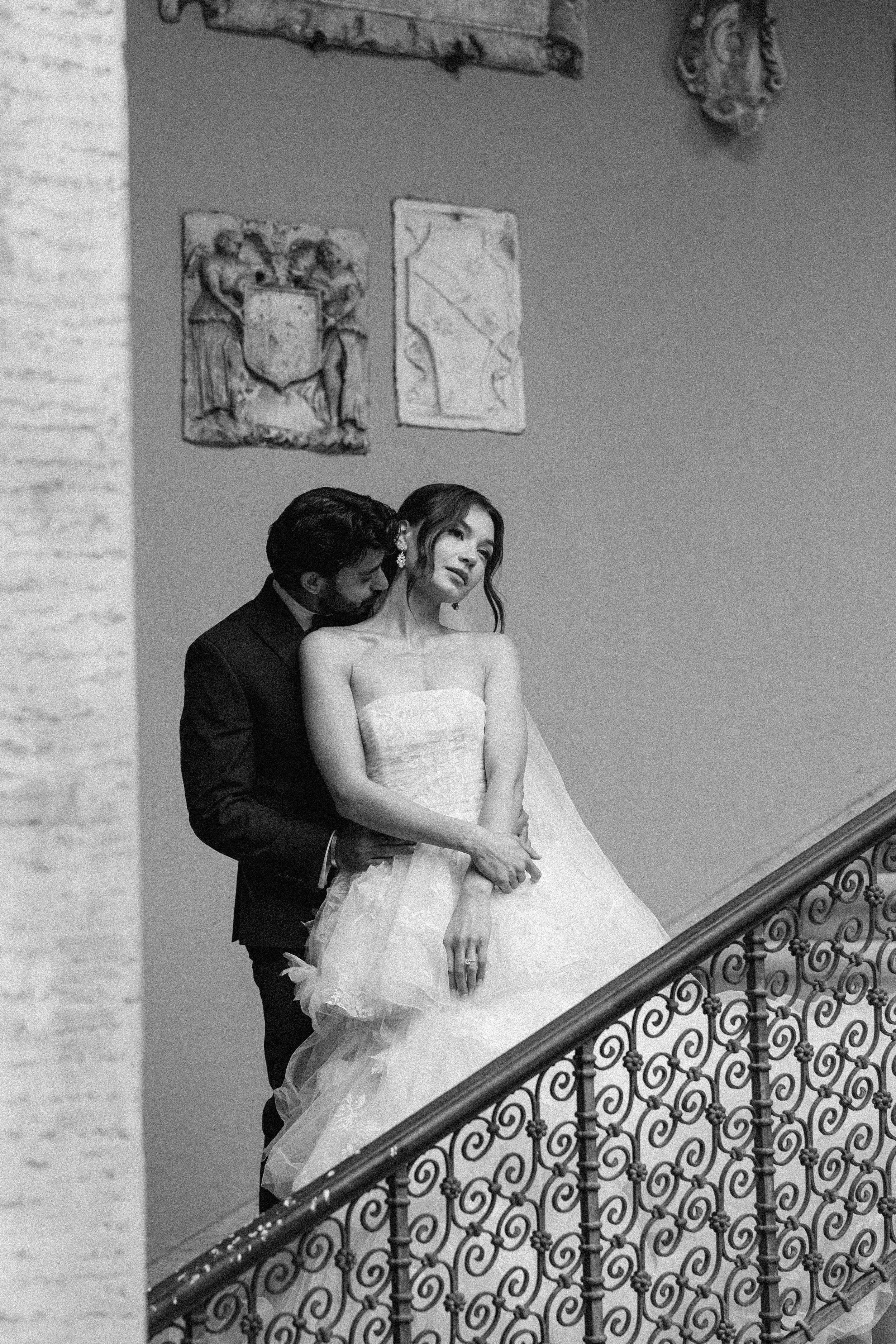 A romantic black and white wedding photograph of a couple on an elegant staircase with ornate railings.