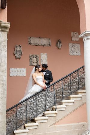 A couple in wedding attire share an intimate moment on an ornate staircase with stone reliefs and a pink wall backdrop.