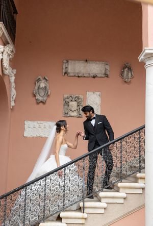 Couple in formal wedding attire pose together on ornate staircase with decorative railing against terracotta colored wall.