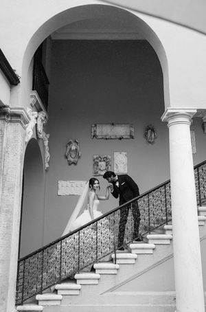 A romantic couple shares an intimate moment on an elegant staircase with white columns and ornate railings.