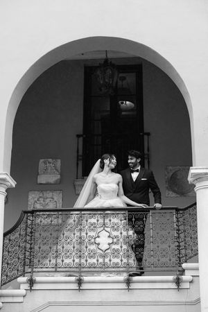 A romantic black and white wedding photo of a couple standing on an ornate balcony under an arched doorway.