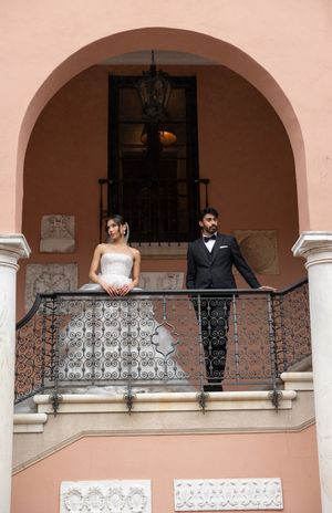 A couple in formal wedding attire pose on an ornate balcony with a pink stucco wall and wrought iron railing.