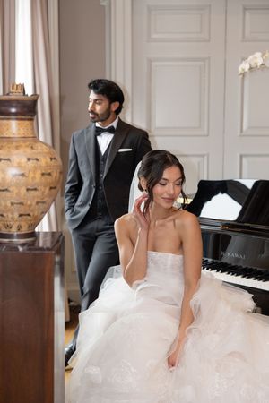 Elegant bride in white gown sits by piano while groom in tuxedo stands behind in luxurious interior.