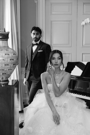 Elegant couple poses in formal attire near a grand piano and decorative vase in a bright white room.
