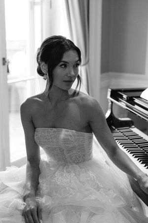 Black and white portrait of a bride in a strapless lace gown standing beside a piano.