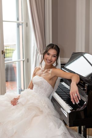A bride in a white strapless wedding gown poses elegantly while seated at a grand piano near a window.