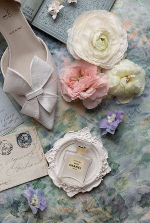 Wedding accessories including blush shoes, roses, and a Chanel perfume bottle arranged on vintage paper.