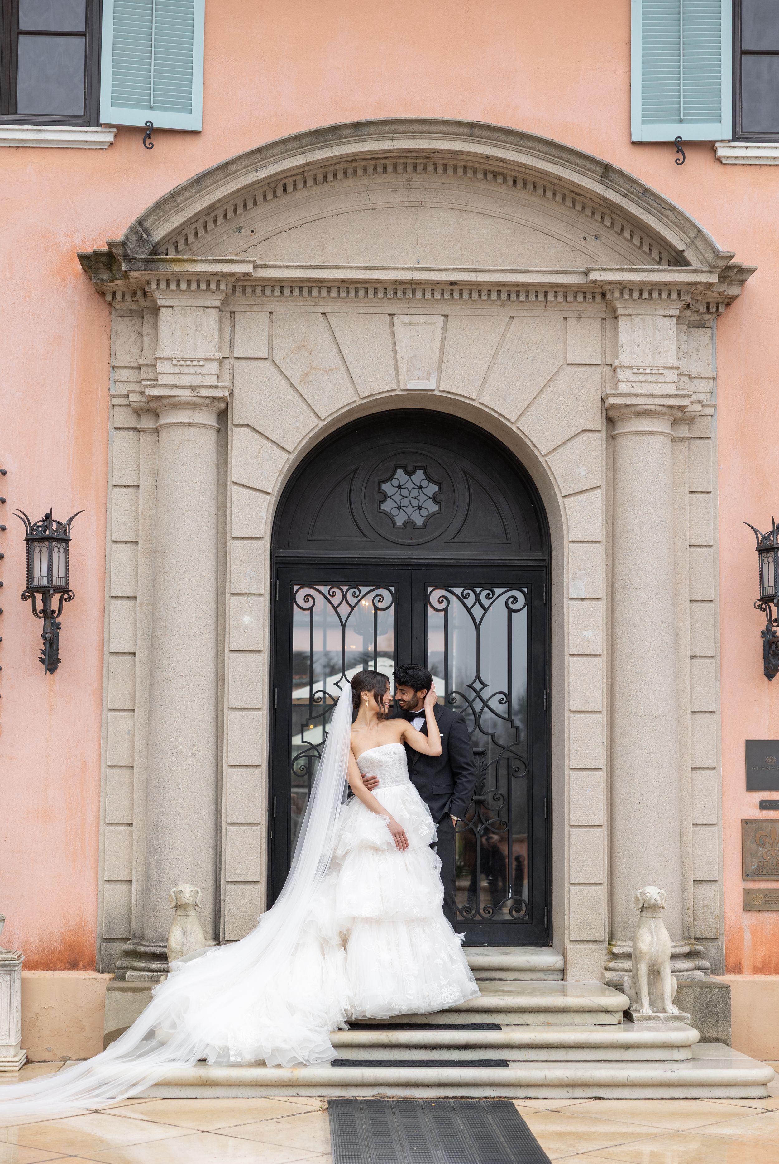 A bride in a white flowing gown poses in front of an elegant arched doorway with ornate stonework and peach-colored walls.