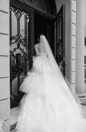 A wedding gown with a flowing cathedral veil seen from behind entering through ornate wrought iron doors.