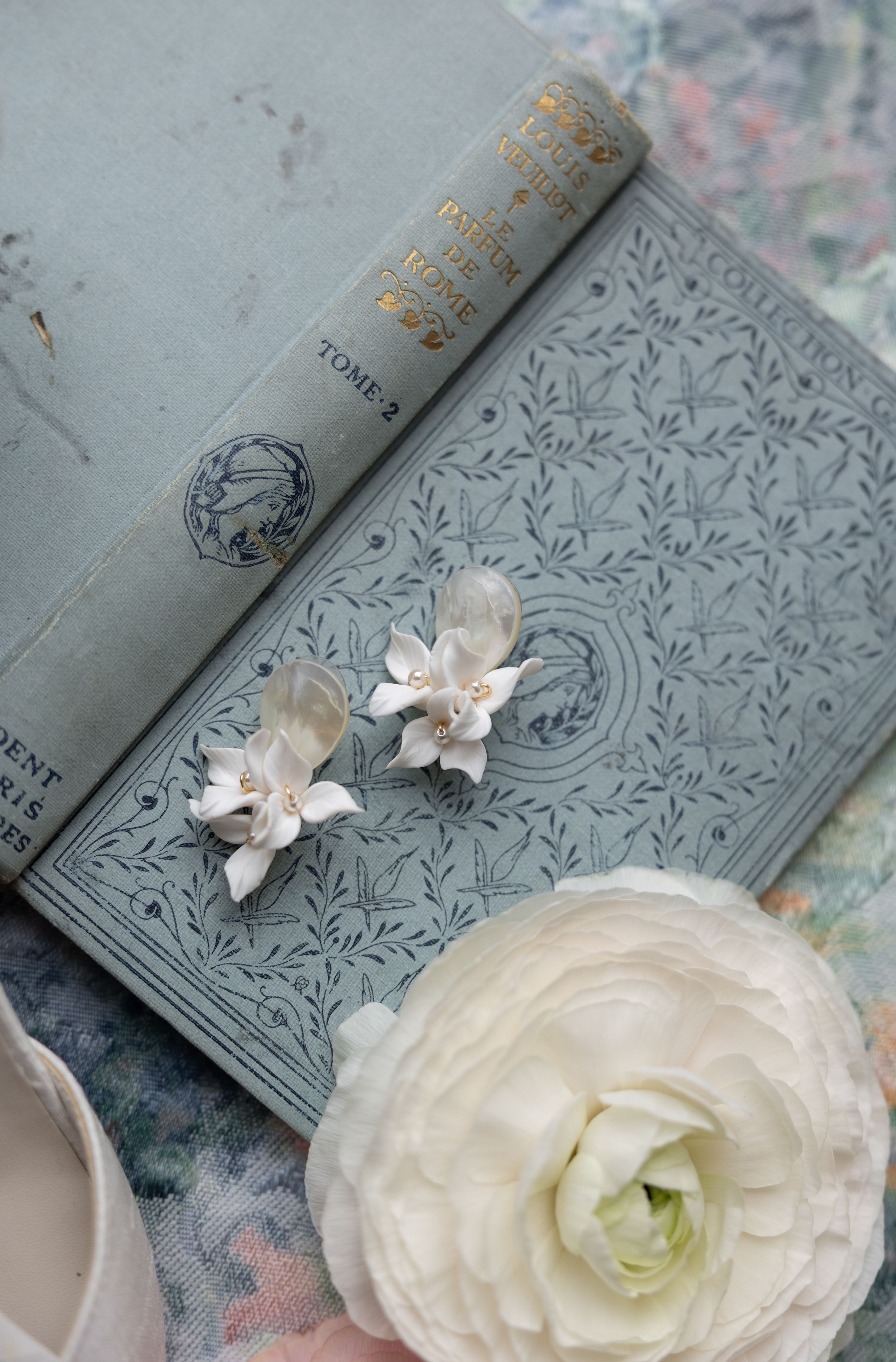 A vintage gray book with ornate cover design displayed with delicate white flowers and a cream-colored ranunculus bloom.