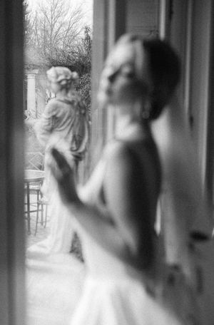 A dreamy black and white photograph shows figures in elegant white dresses standing by a window with soft natural light.