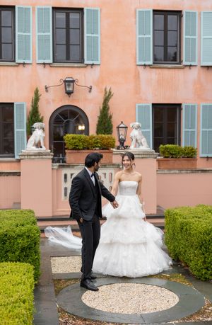 A couple in wedding attire standing in front of a peach-colored mansion with decorative statues.
