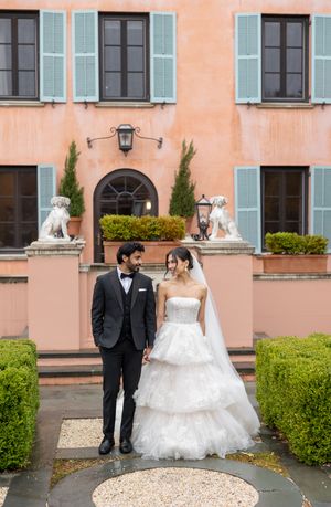 A couple stands in wedding attire in front of a peach-colored European villa with blue shutters and decorative stone lions.