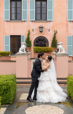 Wedding couple embracing in front of a peach-colored mansion with blue shutters and lion statues.
