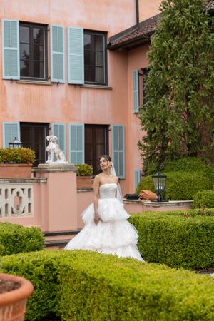 A bride in a white gown poses in a manicured garden with peach-colored villa and green shutters.